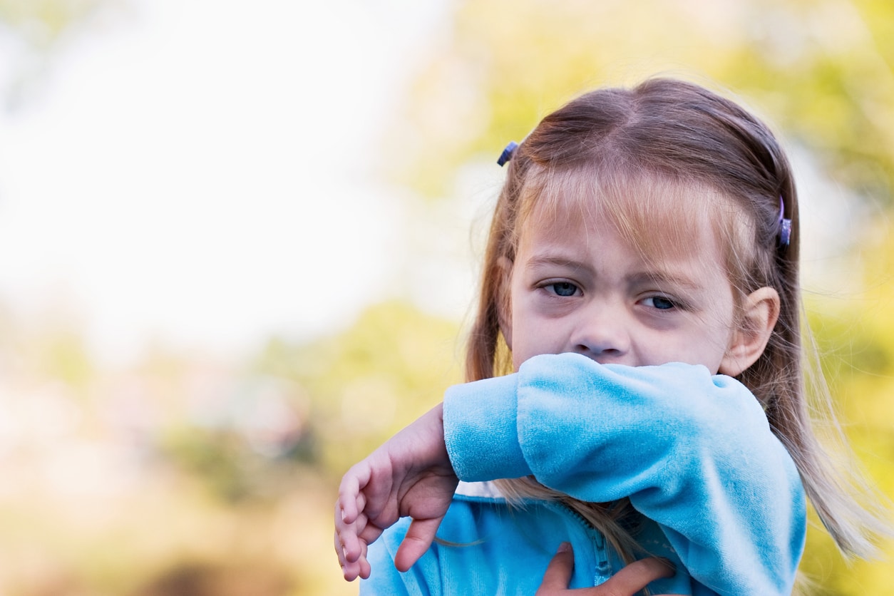 A little girl coughing into her arm while outside.