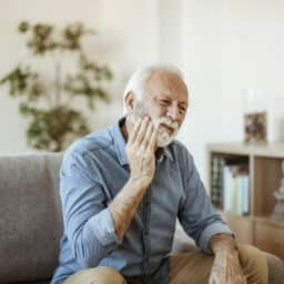 Senior man with toothache sitting on the couch holding his jaw